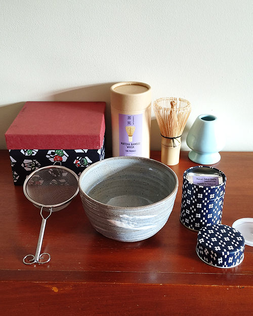 Complete Gokujo Matcha Tea Set displayed on a wooden table, featuring ceremonial-grade matcha in a tin, bamboo scoop, bamboo whisk, whisk stand, tea sifter, and a handcrafted Kiyomizu-ware matcha bowl with gray, white, and blue brushstroke accents.. Gift ideas from j-okini.com in Malta