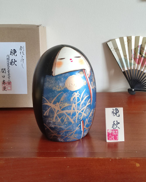 A handcrafted Japanese Kokeshi doll named 'Banshu' by Sekiguchi Toua, standing 16.5 cm tall, featuring a deep blue kimono with gold-painted pampas grass, a moon, and subtle glitters for a shimmering effect, symbolizing late autumn. Available at j-okini.com in Malta