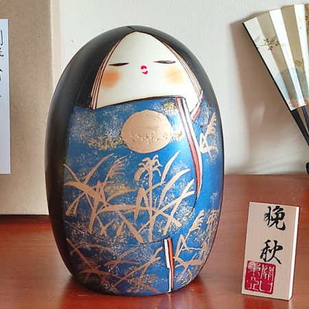 A handcrafted Japanese Kokeshi doll named 'Banshu' by Sekiguchi Toua, standing 16.5 cm tall, featuring a deep blue kimono with gold-painted pampas grass, a moon, and subtle glitters for a shimmering effect, symbolizing late autumn. Available at j-okini.com in Malta