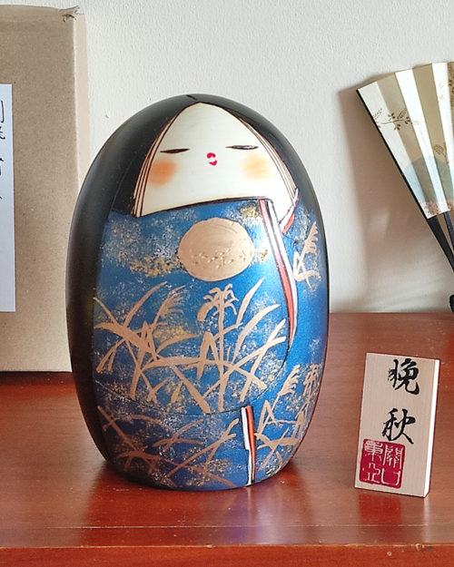 A handcrafted Japanese Kokeshi doll named 'Banshu' by Sekiguchi Toua, standing 16.5 cm tall, featuring a deep blue kimono with gold-painted pampas grass, a moon, and subtle glitters for a shimmering effect, symbolizing late autumn. Available at j-okini.com in Malta