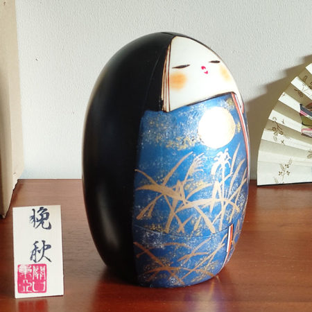 A handcrafted Japanese Kokeshi doll named 'Banshu' by Sekiguchi Toua, standing 16.5 cm tall, featuring a deep blue kimono with gold-painted pampas grass, a moon, and subtle glitters for a shimmering effect, symbolizing late autumn. Available at j-okini.com in Malta