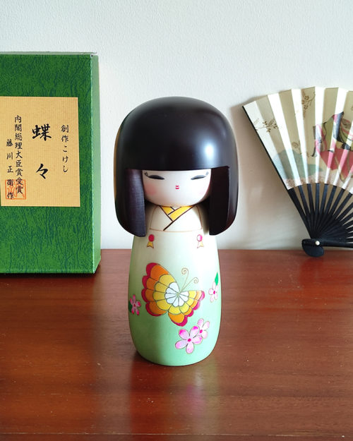 Handcrafted Japanese Kokeshi doll 'Chouchou' by artist Fujikawa Shoei, featuring a light green Michiyuki kimono jacket with a butterfly and pink floral design. Available at j-okini.com in Malta