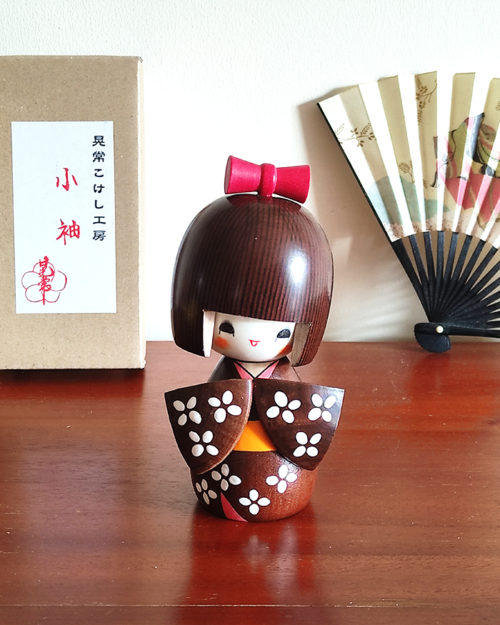 Handcrafted Japanese Kokeshi doll 'Kosode' by artist Kojo, wearing a chic brown kimono with a soft pink collar and a vibrant yellow obi. Available at j-okini.com in Malta