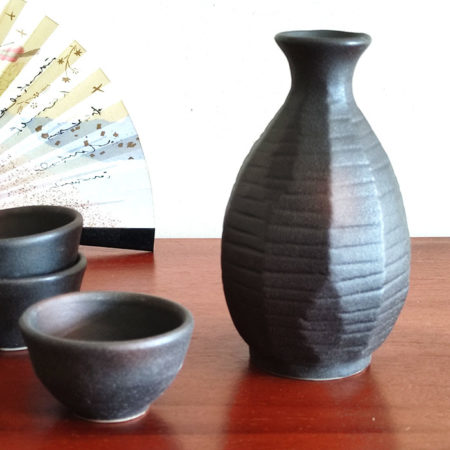 A Sogi Gata Bizen Sake Set featuring a textured black tokkuri bottle with a scraped design and four matching cups, displayed on a wooden surface with a Japanese folding fan in the background. Made in Japan. Available at j-okini.com in Malta