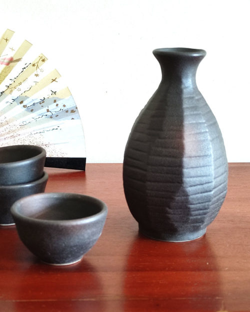 A Sogi Gata Bizen Sake Set featuring a textured black tokkuri bottle with a scraped design and four matching cups, displayed on a wooden surface with a Japanese folding fan in the background. Made in Japan. Available at j-okini.com in Malta