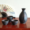 A Sogi Gata Bizen Sake Set featuring a textured black tokkuri bottle with a scraped design and four matching cups, displayed on a wooden surface with a Japanese folding fan in the background. Made in Japan. Available at j-okini.com in Malta