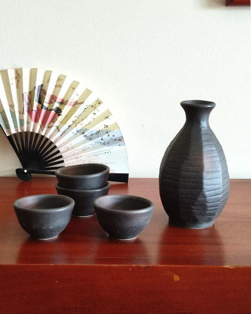 A Sogi Gata Bizen Sake Set featuring a textured black tokkuri bottle with a scraped design and four matching cups, displayed on a wooden surface with a Japanese folding fan in the background. Made in Japan. Available at j-okini.com in Malta