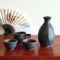 A Sogi Gata Bizen Sake Set featuring a textured black tokkuri bottle with a scraped design and four matching cups, displayed on a wooden surface with a Japanese folding fan in the background. Made in Japan. Available at j-okini.com in Malta