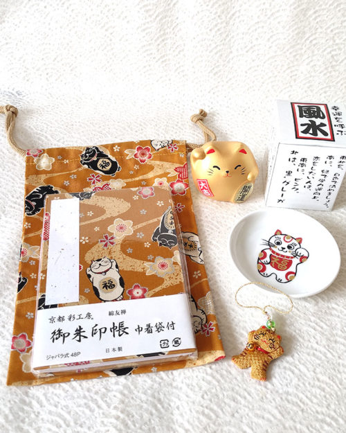 Lucky Cat Blessings Set featuring a golden brown Japanese Goshuincho accordion notebook with a matching drawstring pouch, a white porcelain Maneki-neko small dish, a golden Banko Ware Feng Shui Maneki-neko ceramic figurine, and a handmade Chirimen Cats’ Netsuke strap. A charming collection of Japanese lucky cat-themed items, perfect for gifts or personal good fortune. Made in Japan. Available at j-okini.com in Malta