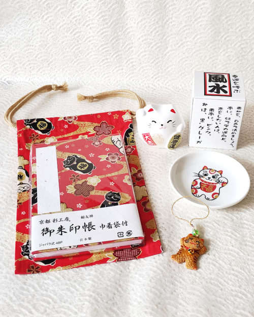 Lucky Cat Blessings Set featuring a red Japanese Goshuincho accordion notebook with a matching drawstring pouch, a white porcelain Maneki-neko small dish, a Banko Ware Feng Shui white Maneki-neko ceramic figurine, and a handmade Chirimen Cats’ Netsuke strap. A charming collection of Japanese lucky cat-themed items, perfect for gifts or personal good fortune. Made in Japan. Available at j-okini.com in Malta.